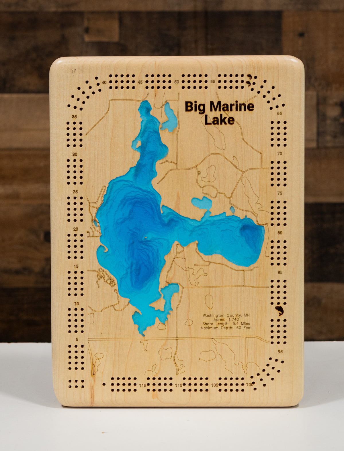 A wooden board game designed as a map of Big Marine Lake, Michigan, featuring a blue resin lake design and engraved playing pieces holes around the border, set against a dark wood background.