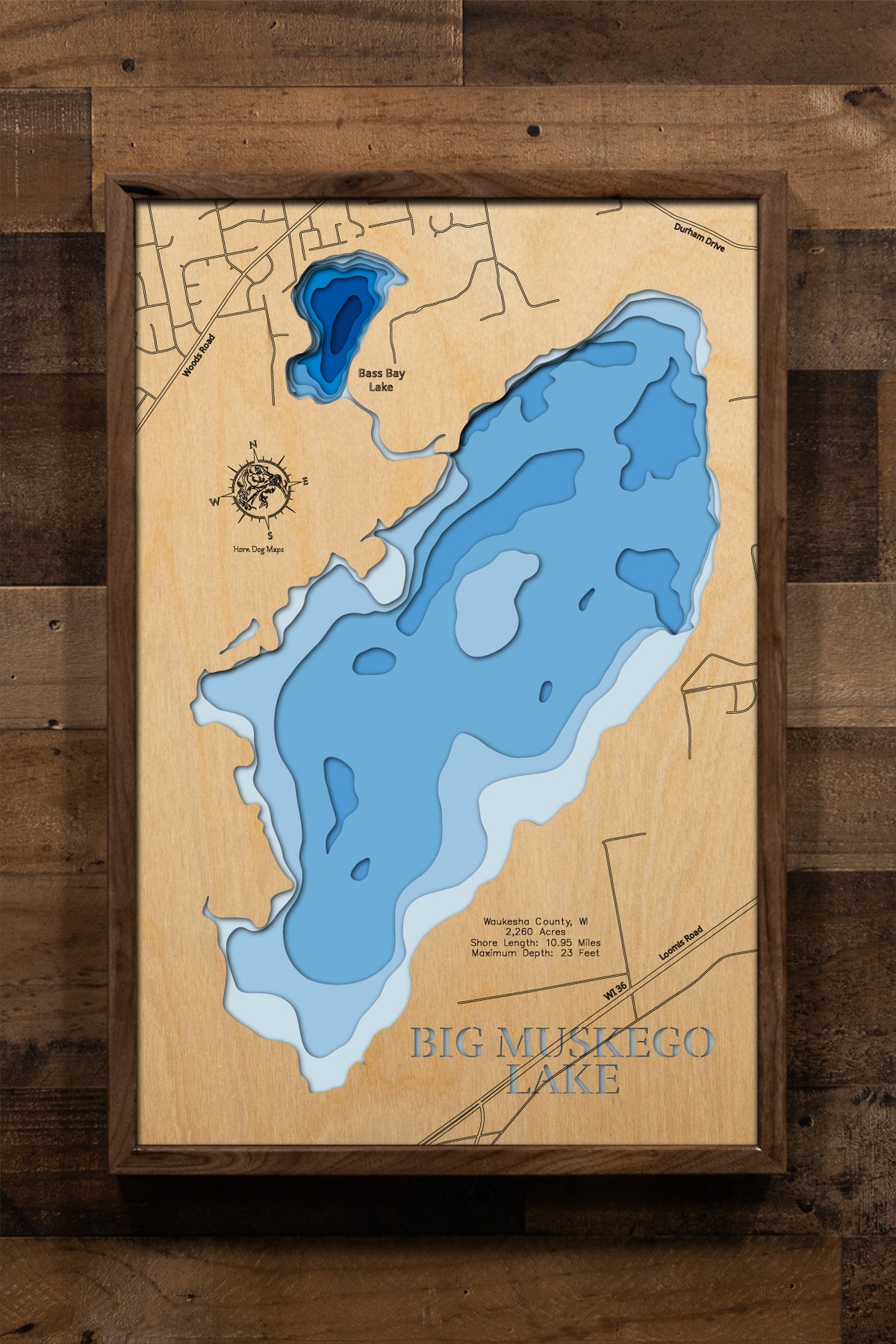A highly detailed, laser-cut, and multi-layered wooden map of Big Muskego Lake, a large natural lake in Waukesha County, Wisconsin. The map shows the lake's distinct shape and its 10.95-mile shoreline. The varying depths of the lake, reaching a maximum of 23 feet, are represented by different layers of wood.