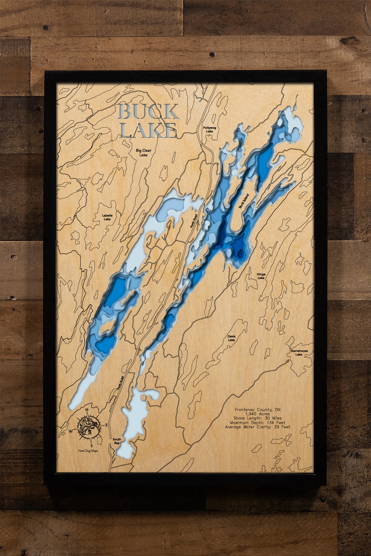A highly detailed, laser-cut, and multi-layered wooden map of Buck Lake in Frontenac County, Ontario. The map showcases the lake's 1,940-acre surface divided into distinctive North and South Branches connected by a culvert, depth contours illustrating the dramatic descent to 134 feet, multiple rocky islands and shoals, and 30 miles of Canadian Shield granite shoreline.