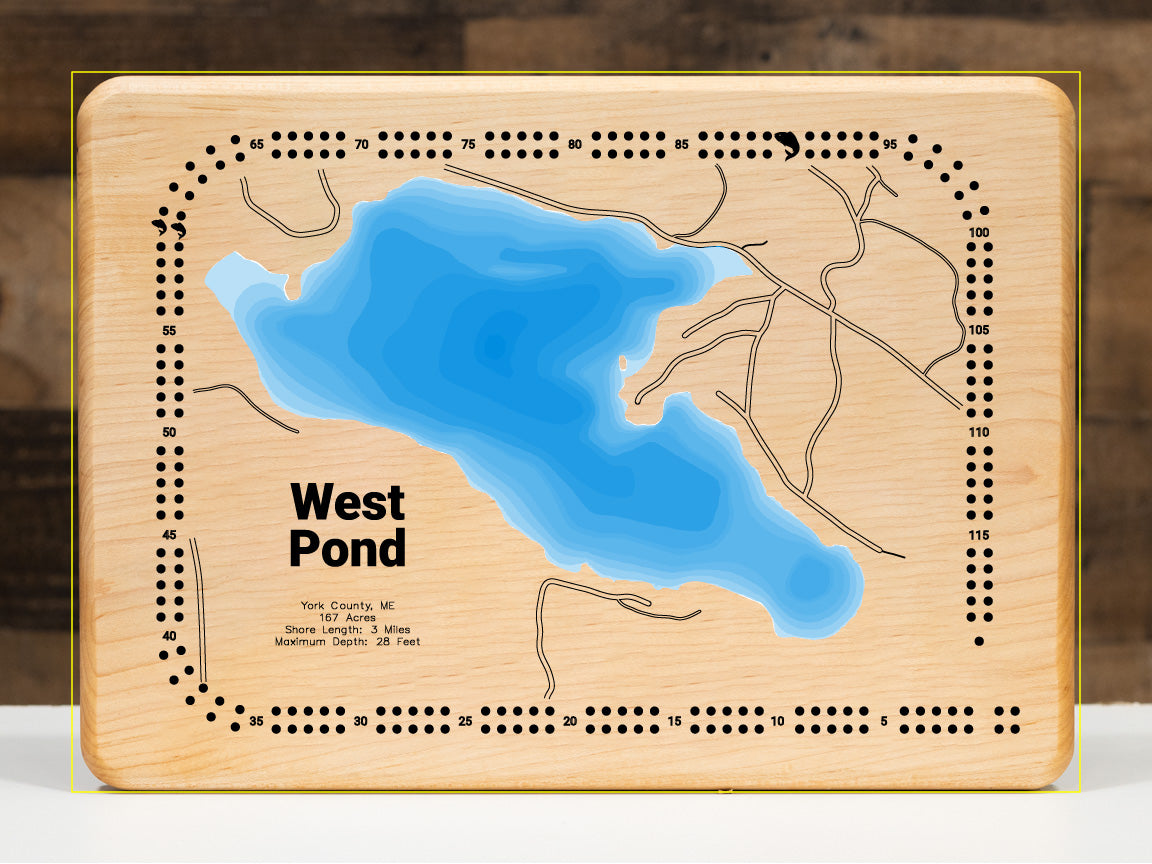 A meticulously detailed, laser-cut, multi-layered wooden topographical map of West Pond in York County, Maine, showcasing the intimate 167-acre lake tucked in the foothills of the White Mountains with depth contours from the 10-foot average to the 28-foot maximum depth. The natural wood top layer frames three miles of shoreline with the outlet dam and pristine waters, while graduated blue layers create a gentle visual depth gradient.