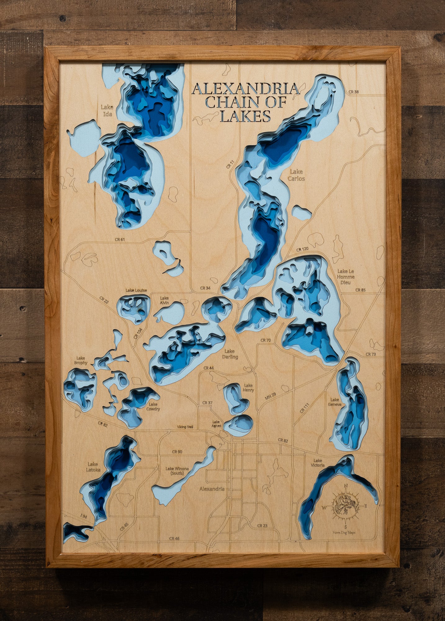 A highly detailed, laser-cut, and multi-layered wooden map of the Alexandria Chain of Lakes in Douglas County, Minnesota. The map shows the distinct shapes of its major lakes and the extensive, interconnected shoreline of 55 miles. The varying depths of the chain, reaching a maximum of 85 feet, are represented by different layers of wood.
