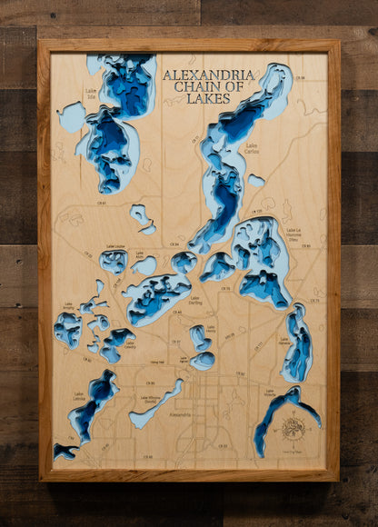 A highly detailed, laser-cut, and multi-layered wooden map of the Alexandria Chain of Lakes in Douglas County, Minnesota. The map shows the distinct shapes of its major lakes and the extensive, interconnected shoreline of 55 miles. The varying depths of the chain, reaching a maximum of 85 feet, are represented by different layers of wood.