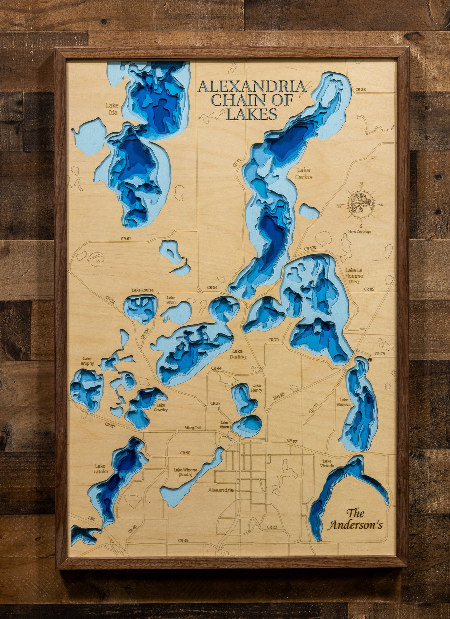 A highly detailed, laser-cut, and multi-layered wooden map of the Alexandria Chain of Lakes in Douglas County, Minnesota. The map shows the distinct shapes of its major lakes and the extensive, interconnected shoreline of 55 miles. The varying depths of the chain, reaching a maximum of 85 feet, are represented by different layers of wood.