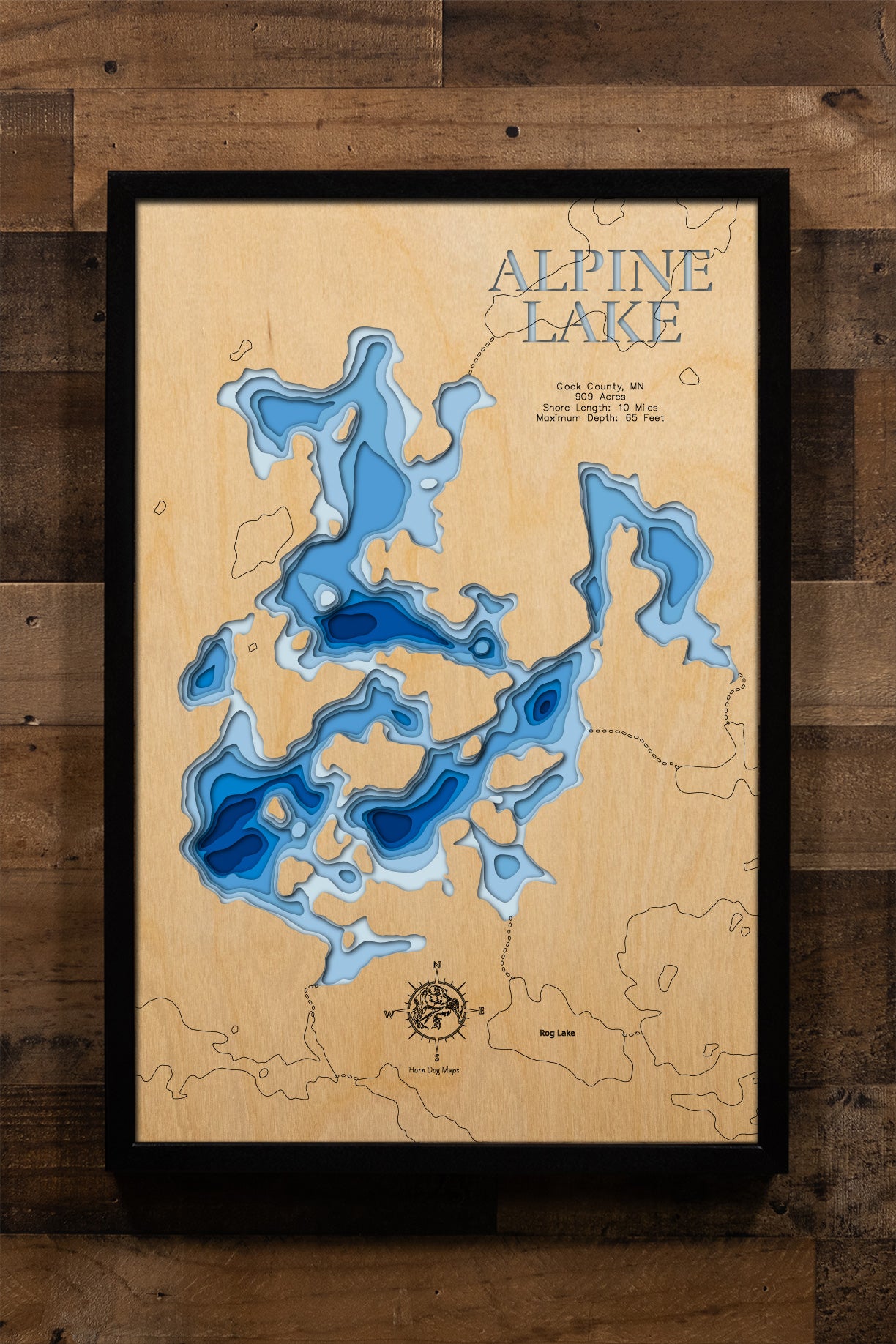 A meticulously detailed, laser-cut, multi-layered wooden topographical map of Alpine Lake in Cook County, Minnesota, showcasing the 909-acre wilderness lake within Superior National Forest with depth contours from moderate depths to the 65-foot maximum. The natural wood top layer frames approximately 10 miles of pristine shoreline near the Boundary Waters Canoe Area Wilderness, while graduated blue layers create a striking visual depth gradient.