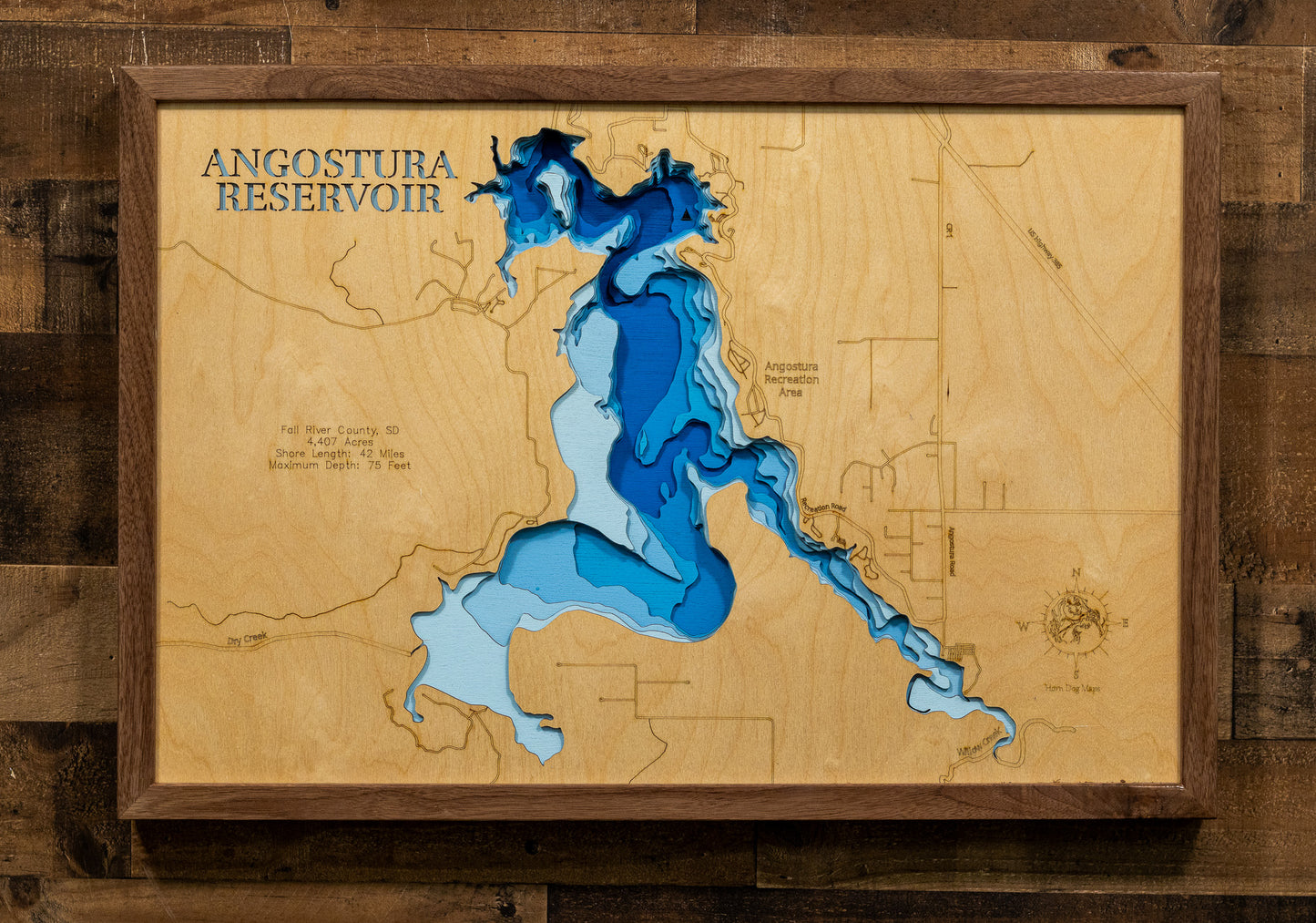 A framed wooden depth map of Angostura Reservoir, featuring layered blue and light blue wood sections representing water depth over a natural wood grain background, mounted on a dark wood plank wall.