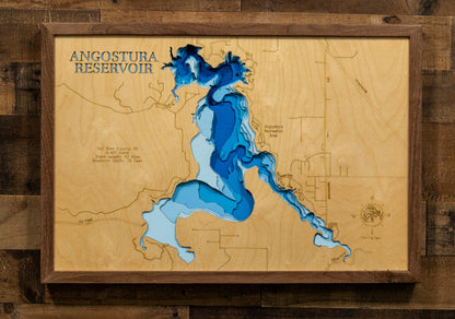 A framed wooden depth map of Angostura Reservoir, featuring layered blue and light blue wood sections representing water depth over a natural wood grain background, mounted on a dark wood plank wall.