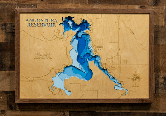A framed wooden depth map of Angostura Reservoir, featuring layered blue and light blue wood sections representing water depth over a natural wood grain background, mounted on a dark wood plank wall.