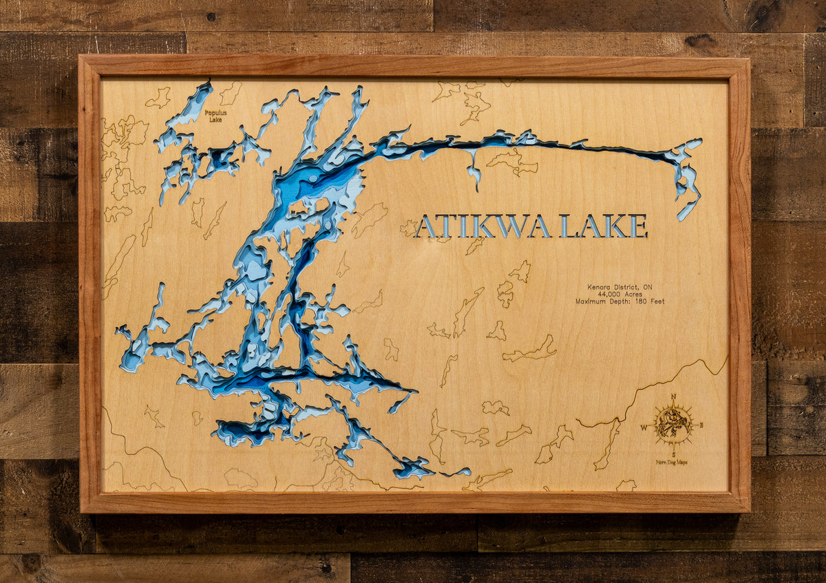 A framed wooden Atikwa Lake map wall art piece, featuring a light natural wood background, blue lake details, and laser-engraved text and coastline, mounted on a dark wood plank wall.