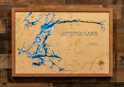 A framed wooden Atikwa Lake map wall art piece, featuring a light natural wood background, blue lake details, and laser-engraved text and coastline, mounted on a dark wood plank wall.