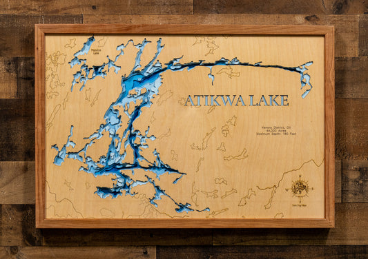 A framed wooden Atikwa Lake map wall art piece, featuring a light natural wood background, blue lake details, and laser-engraved text and coastline, mounted on a dark wood plank wall.