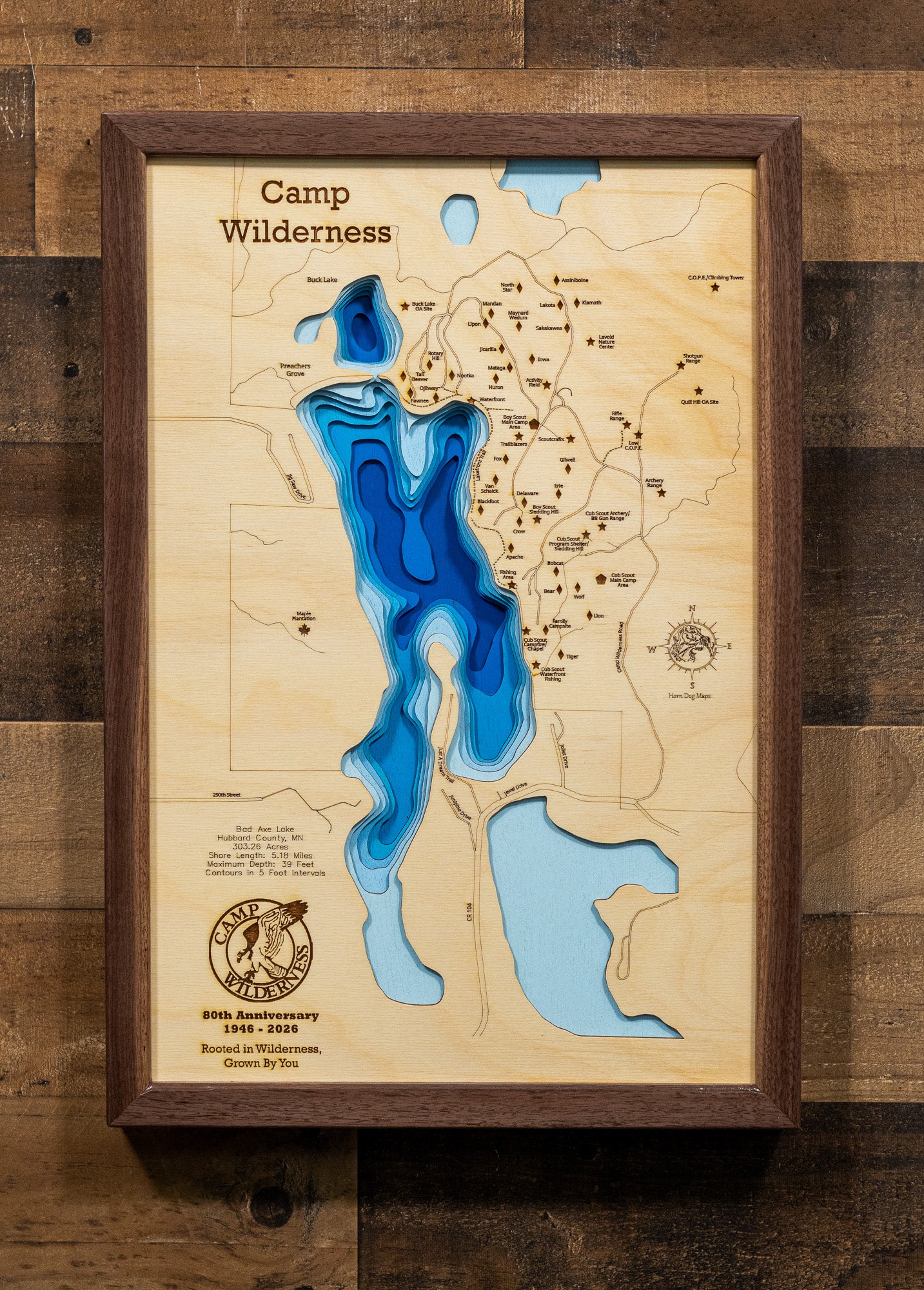 A precision laser-cut, multi-layered wooden map of Bad Axe Lake in Hubbard County, Minnesota — home to the legendary Camp Wilderness Scout Camp. The natural wood top layer traces the lake's pristine, protected shoreline in fine detail, while a light-to-dark blue gradient highlights the water's depth and dimension. A meaningful and rustic statement piece for a lodge, troop room, or Scout family home.