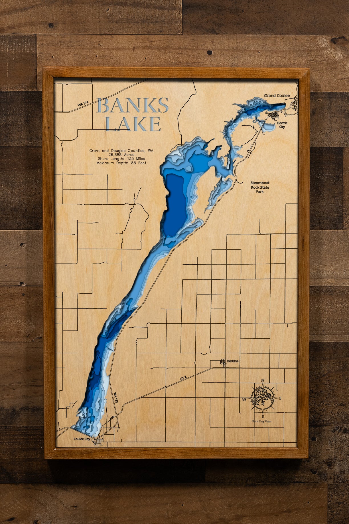 A precision laser-cut, multi-layered wooden map of Banks Lake in Grant and Douglas Counties, Washington. The natural wood top layer captures 135 miles of dramatic basalt-cliff shoreline and the lake's sweeping 27-mile coulee in fine detail, while a light-to-dark blue gradient highlights the water's depth and dimension. A stunning, rustic statement piece for any Pacific Northwest cabin, lake home, or outdoor enthusiast's space.