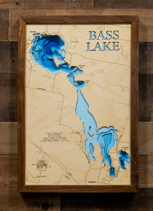 A framed Bass Lake wooden map art piece with layered blue and light blue depth details and a dark wood frame, displayed against a rustic dark wood plank wall.