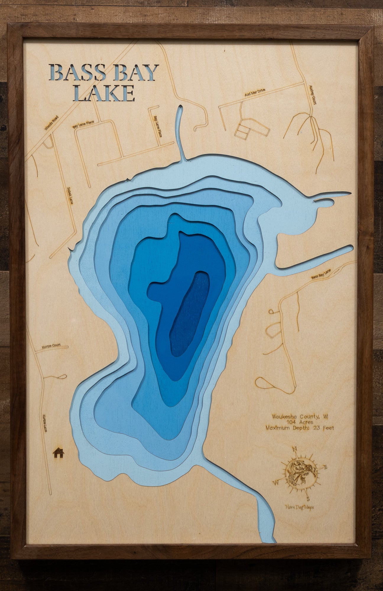 A highly detailed, laser-cut, and multi-layered wooden map of Bass Bay, a natural lake in Waukesha County, Wisconsin. The map shows the lake's distinct shape and its connection to the larger Big Muskego Lake. The varying depths of the lake, reaching a maximum of 23 feet, are represented by different layers of wood.