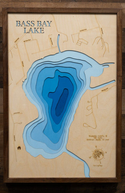 A highly detailed, laser-cut, and multi-layered wooden map of Bass Bay, a natural lake in Waukesha County, Wisconsin. The map shows the lake's distinct shape and its connection to the larger Big Muskego Lake. The varying depths of the lake, reaching a maximum of 23 feet, are represented by different layers of wood.