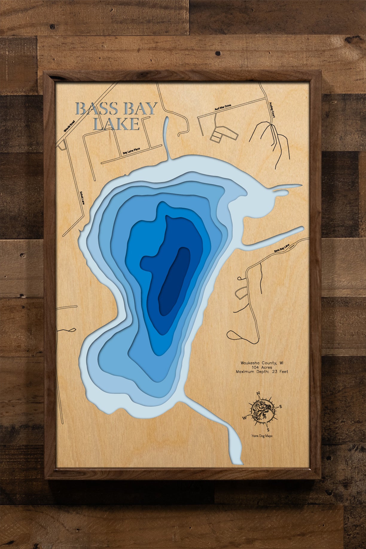 A highly detailed, laser-cut, and multi-layered wooden map of Bass Bay, a natural lake in Waukesha County, Wisconsin. The map shows the lake's distinct shape and its connection to the larger Big Muskego Lake. The varying depths of the lake, reaching a maximum of 23 feet, are represented by different layers of wood.
