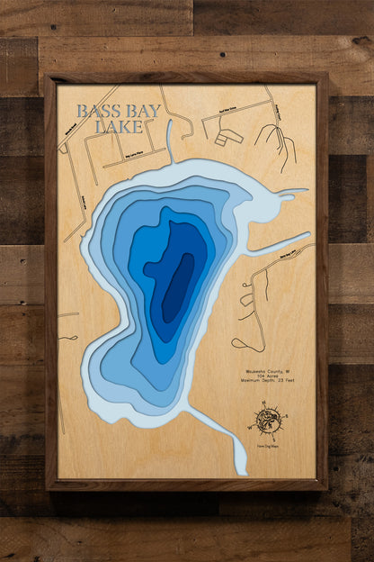 A highly detailed, laser-cut, and multi-layered wooden map of Bass Bay, a natural lake in Waukesha County, Wisconsin. The map shows the lake's distinct shape and its connection to the larger Big Muskego Lake. The varying depths of the lake, reaching a maximum of 23 feet, are represented by different layers of wood.