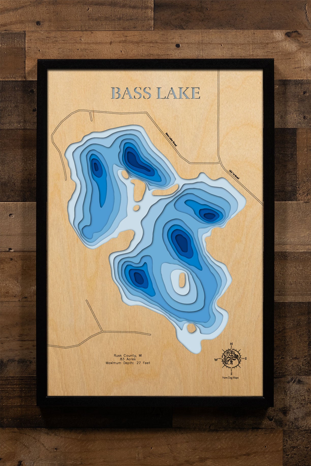 A precision laser-cut, multi-layered wooden map of Bass Lake in Rusk County, Wisconsin, an 83-acre Northwoods lake near Strickland holding Musky, Walleye, Trout, Bass, and Pike. The natural wood top layer captures the lake's natural shoreline in fine detail, while a light-to-dark blue gradient conveys its 27-foot depth. A handcrafted statement piece for any Wisconsin lake cabin, home, or outdoor enthusiast's wall.