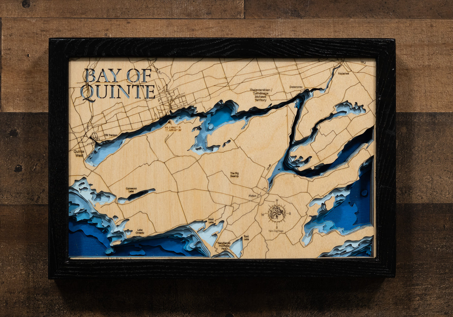 A laser-cut, layered wooden map of the Bay of Quinte in Ontario, Canada. The distinct, sinuous shape of this long bay, which connects to Lake Ontario, is clearly visible as it winds through the landscape of southeastern Ontario. The varying depths of the bay, reaching a maximum of 30 meters, are represented by different layers of wood, illustrating its impressive underwater topography. The location of key communities and landmarks are also visible on the map.