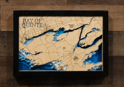 A laser-cut, layered wooden map of the Bay of Quinte in Ontario, Canada. The distinct, sinuous shape of this long bay, which connects to Lake Ontario, is clearly visible as it winds through the landscape of southeastern Ontario. The varying depths of the bay, reaching a maximum of 30 meters, are represented by different layers of wood, illustrating its impressive underwater topography. The location of key communities and landmarks are also visible on the map.