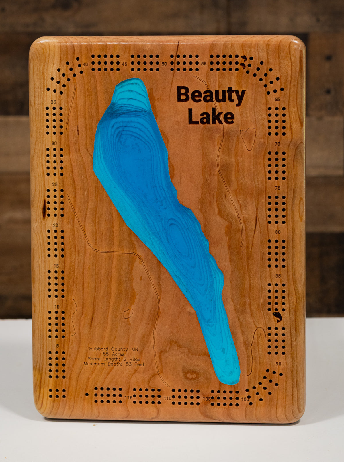 A precision laser-cut, multi-layered wooden map of Beauty Lake in Hubbard County, Minnesota — a pristine 55-acre clear-water lake 19 miles north of Park Rapids with a surprising maximum depth of 53 feet. The natural wood top layer captures 2 miles of pine-fringed shoreline in fine detail, while a light-to-dark blue gradient conveys the lake's remarkable depth and clarity. A handcrafted statement piece for any Minnesota Northwoods cabin, lake home, or outdoor enthusiast's wall.