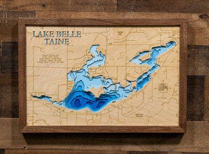 A framed 3D wooden map of Lake Belle Taine, featuring layered wood sections in shades of blue and white to represent the lake's depth, mounted on a rustic dark wood plank wall.