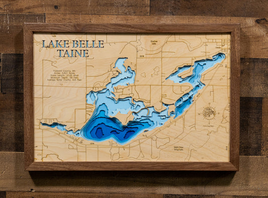A framed 3D wooden map of Lake Belle Taine, featuring layered wood sections in shades of blue and white to represent the lake's depth, mounted on a rustic dark wood plank wall.