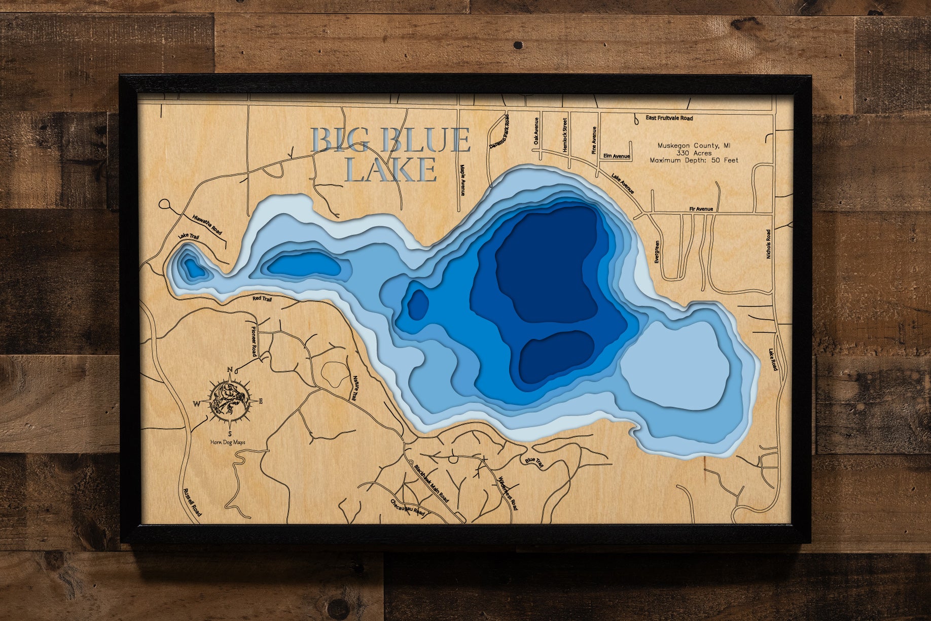 A detailed, laser-cut, multi-layered wooden map of Big Blue Lake in Muskegon County, Michigan, showcasing the 330-acre lake with crystal-clear waters near Whitehall and Montague. The topographic design features natural wood layers highlighting the sandy bottom and surrounding forest landscape, with graduated blue water layers illustrating depths to the 50-foot maximum.