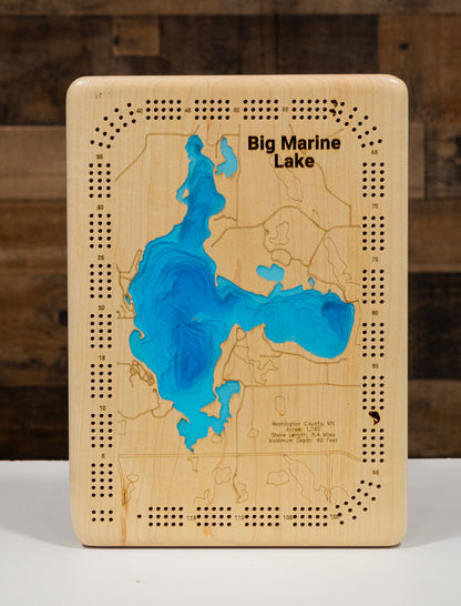 A wooden board game designed as a map of Big Marine Lake, Michigan, featuring a blue resin lake design and engraved playing pieces holes around the border, set against a dark wood background.
