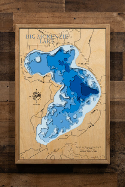 A meticulously detailed, laser-cut, multi-layered wooden topographical map of Big McKenzie Lake in Burnett and Washburn Counties, Wisconsin, showcasing the lake's 1,129-acre surface with intricate depth contours illustrating underwater humps and the transition from shallow bays to the 71-foot maximum depth in the central basin. The natural wood top layer frames 12 miles of precisely rendered shoreline, while graduated blue layers create a striking visual depth gradient.