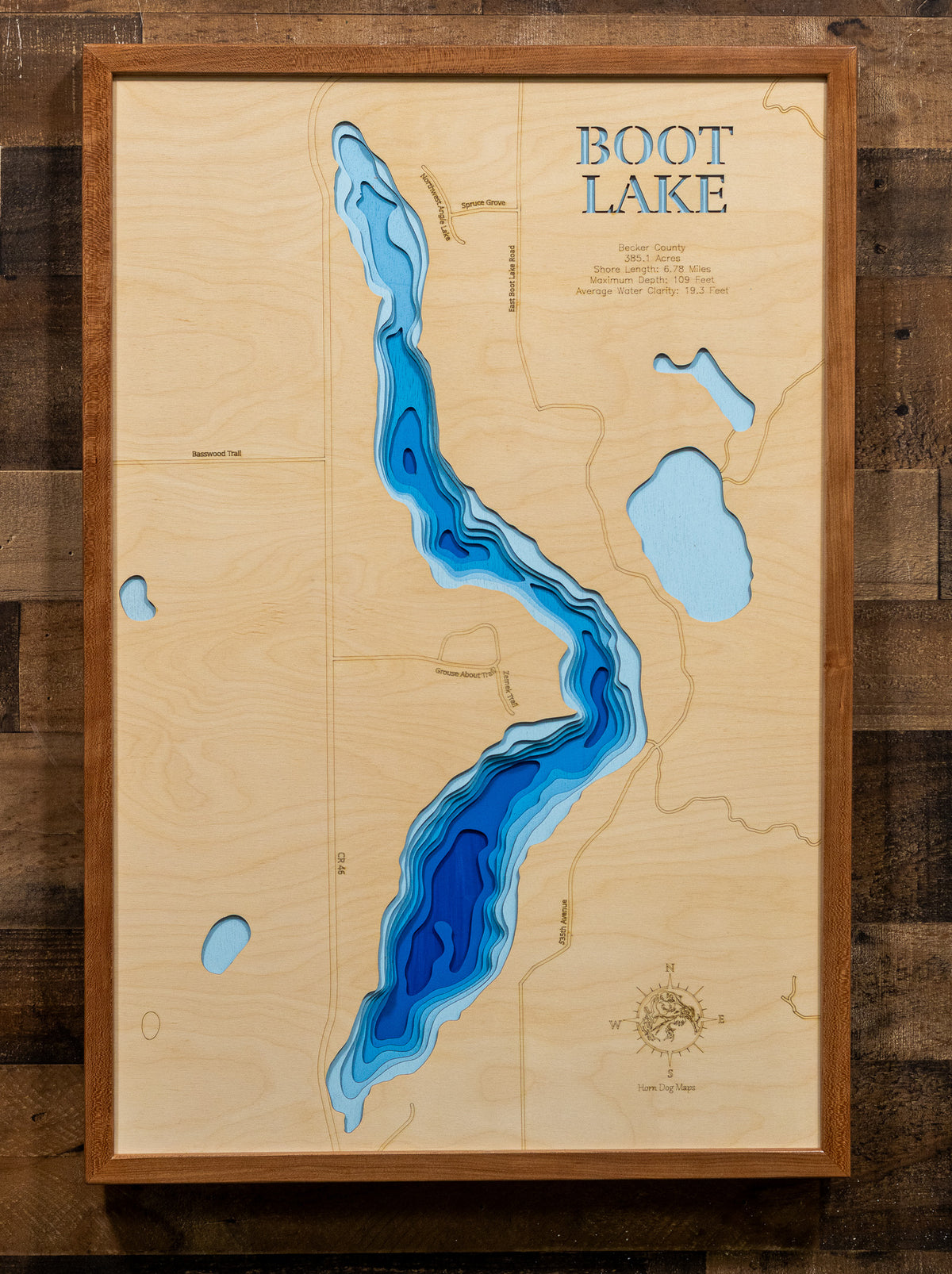 A framed wooden depth map of Boot Lake, featuring layered blue and cyan contours showing water depth on a light wood background, enclosed in a medium brown wood frame and mounted on a dark wood plank wall.