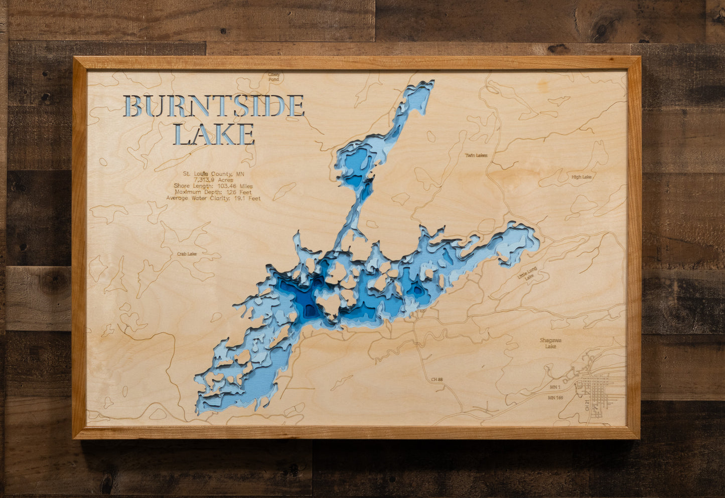 A laser-cut, layered wooden map of Burntside Lake in St. Louis County, Minnesota. The distinct, complex shape of this large natural lake, with its 103.46-mile shoreline and numerous islands, is clearly visible within the forested landscape of northeastern Minnesota. The varying depths of the lake, reaching a remarkable maximum of 126 feet, are represented by different layers of wood, illustrating its impressive underwater topography.