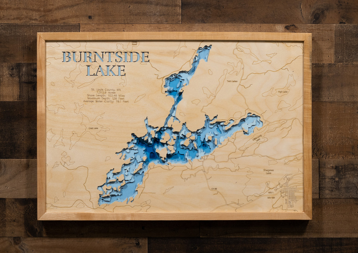 A laser-cut, layered wooden map of Burntside Lake in St. Louis County, Minnesota. The distinct, complex shape of this large natural lake, with its 103.46-mile shoreline and numerous islands, is clearly visible within the forested landscape of northeastern Minnesota. The varying depths of the lake, reaching a remarkable maximum of 126 feet, are represented by different layers of wood, illustrating its impressive underwater topography.