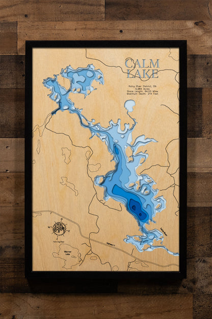 A precision laser-cut, multi-layered wooden map of Calm Lake in Rainy River District, Ontario, the 5,984-acre headwaters of the Seine River Chain near Atikokan with 64 miles of Canadian Shield shoreline and depths exceeding 200 feet. The natural wood top layer captures every dramatic bay and granite point in fine detail, while a light-to-dark blue gradient conveys the lake's remarkable depth. A handcrafted statement piece for any Ontario fishing camp, wilderness cabin, or Quetico-area enthusiast's wall.