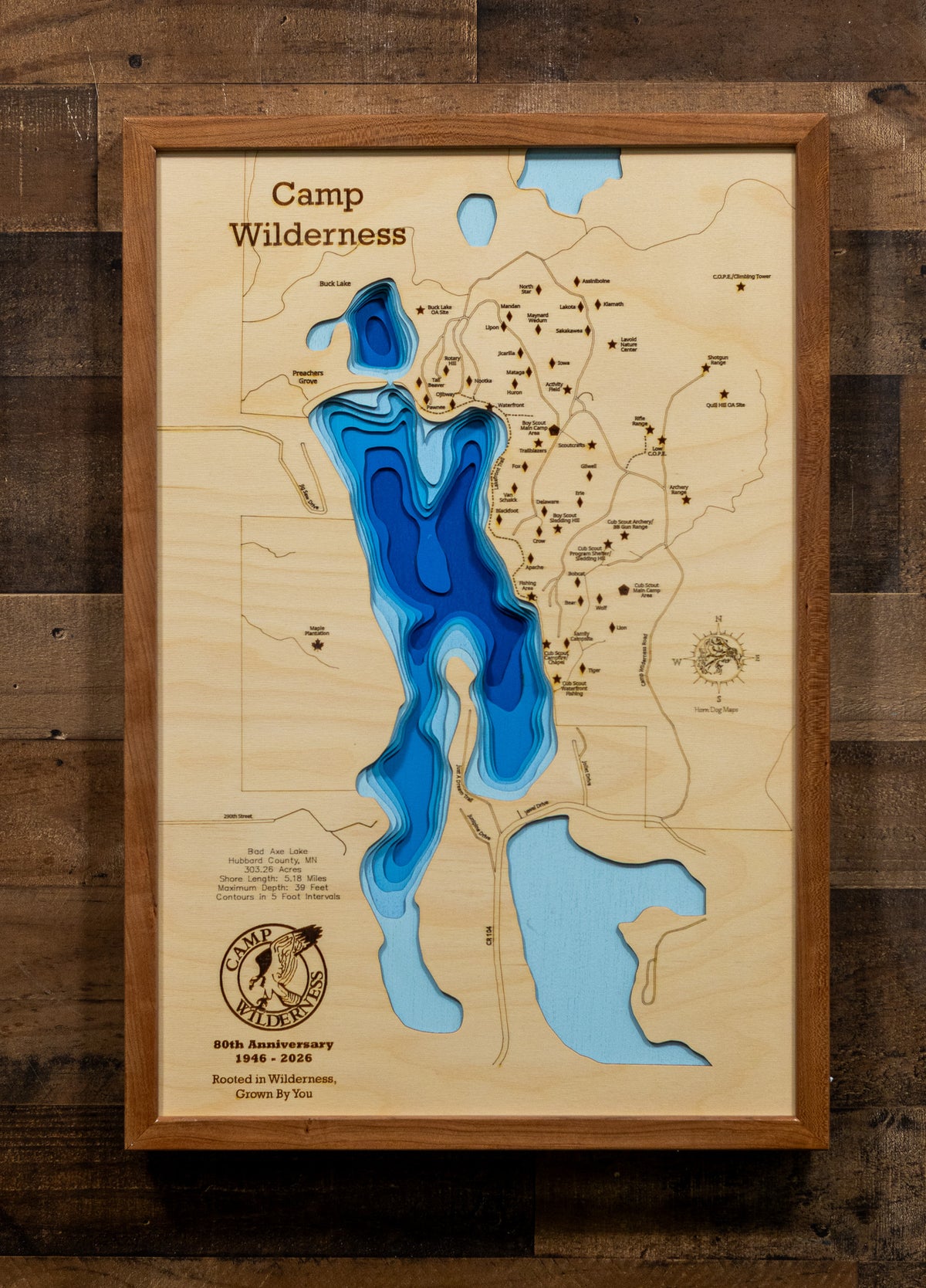 A framed 3D wooden topographic map of Camp Wilderness, featuring layered blue and light blue resin cutouts representing lakes and water features, mounted on a rustic dark wood plank wall.