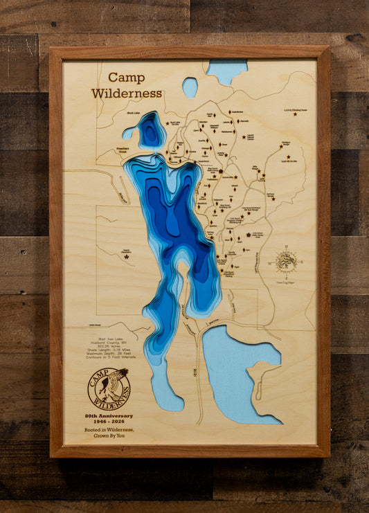A framed 3D wooden topographic map of Camp Wilderness, featuring layered blue and light blue resin cutouts representing lakes and water features, mounted on a rustic dark wood plank wall.