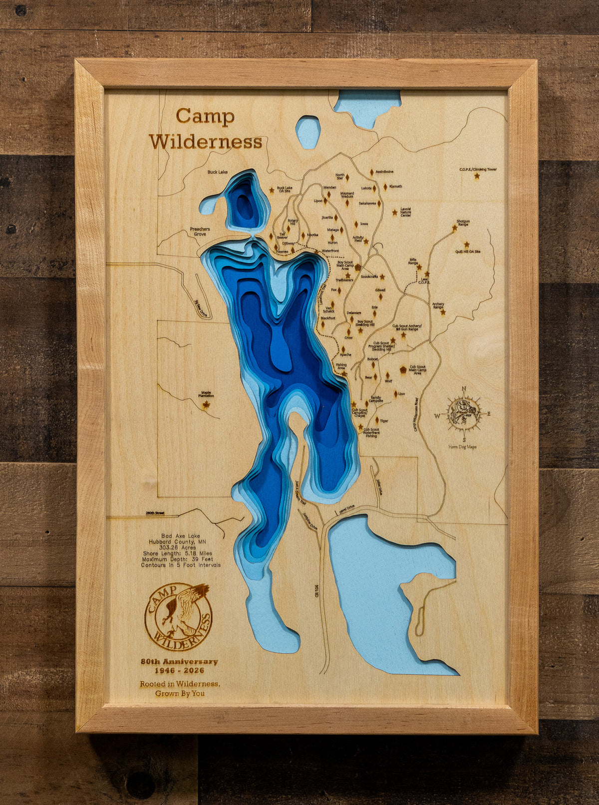 A framed wooden map wall art piece titled "Camp Wilderness" with layered blue and light blue lake shapes, shown hanging on a dark wood plank wall.