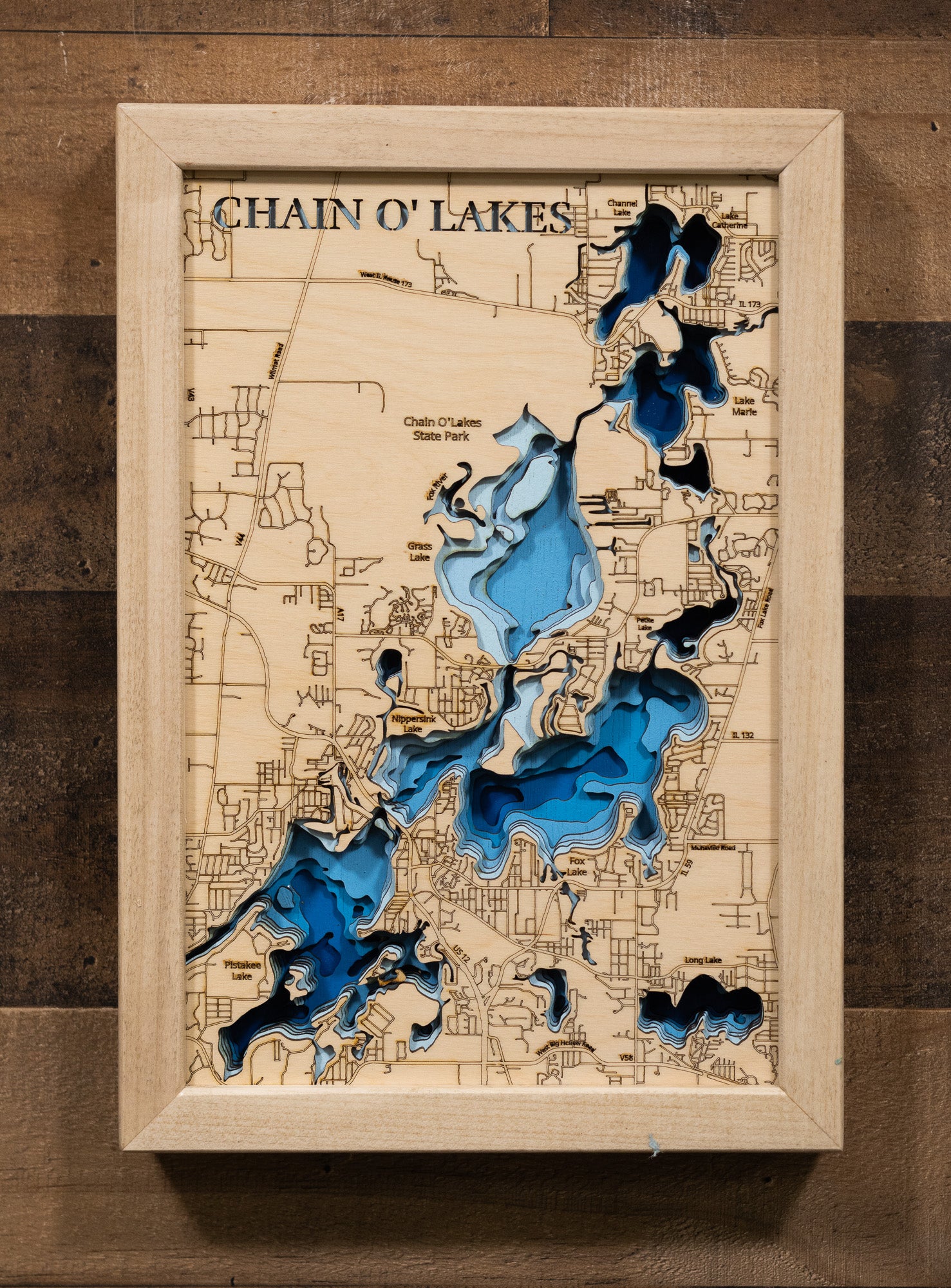 A laser-cut, layered wooden map of the Chain O'Lakes, located in McHenry and Lake Counties, Illinois. The interconnected and intricate shapes of the 15 lakes and connecting waterways are clearly visible within the northeastern Illinois landscape. The varying depths of the individual lakes are represented by different layers of wood, illustrating the underwater topography of this unique system.