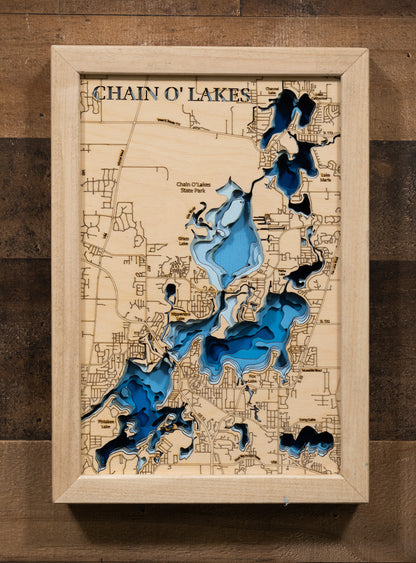 A laser-cut, layered wooden map of the Chain O'Lakes, located in McHenry and Lake Counties, Illinois. The interconnected and intricate shapes of the 15 lakes and connecting waterways are clearly visible within the northeastern Illinois landscape. The varying depths of the individual lakes are represented by different layers of wood, illustrating the underwater topography of this unique system.