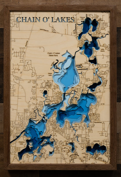A laser-cut, layered wooden map of the Chain O'Lakes, located in McHenry and Lake Counties, Illinois. The interconnected and intricate shapes of the 15 lakes and connecting waterways are clearly visible within the northeastern Illinois landscape. The varying depths of the individual lakes are represented by different layers of wood, illustrating the underwater topography of this unique system.