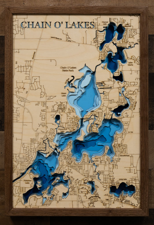 A laser-cut, layered wooden map of the Chain O'Lakes, located in McHenry and Lake Counties, Illinois. The interconnected and intricate shapes of the 15 lakes and connecting waterways are clearly visible within the northeastern Illinois landscape. The varying depths of the individual lakes are represented by different layers of wood, illustrating the underwater topography of this unique system.