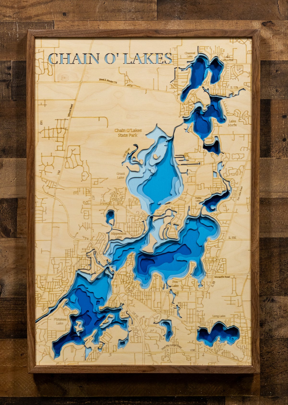 A framed 3D wooden map of Chain O' Lakes, Michigan, featuring layered cutouts in shades of blue to represent water depth, mounted on a rustic dark wood plank wall.