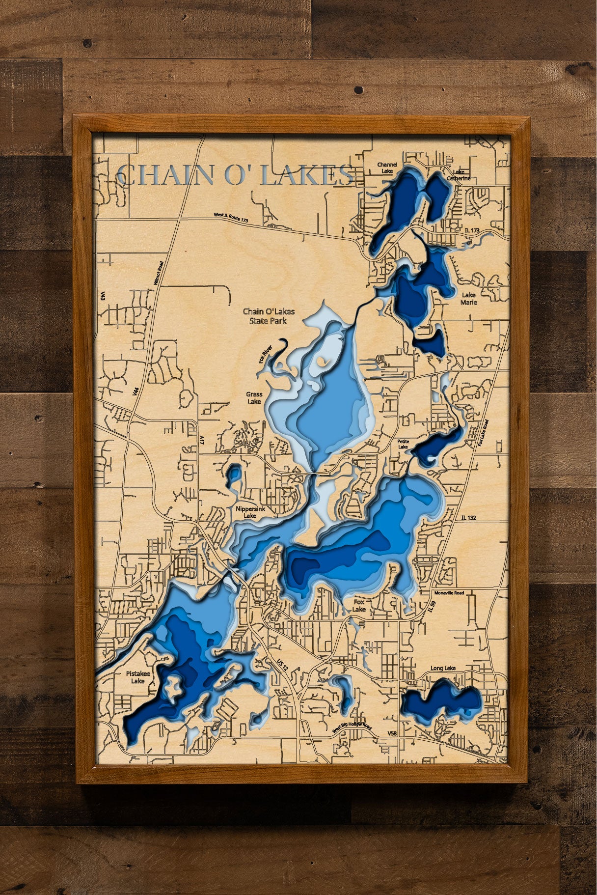 A laser-cut, layered wooden map of the Chain O'Lakes, located in McHenry and Lake Counties, Illinois. The interconnected and intricate shapes of the 15 lakes and connecting waterways are clearly visible within the northeastern Illinois landscape. The varying depths of the individual lakes are represented by different layers of wood, illustrating the underwater topography of this unique system.