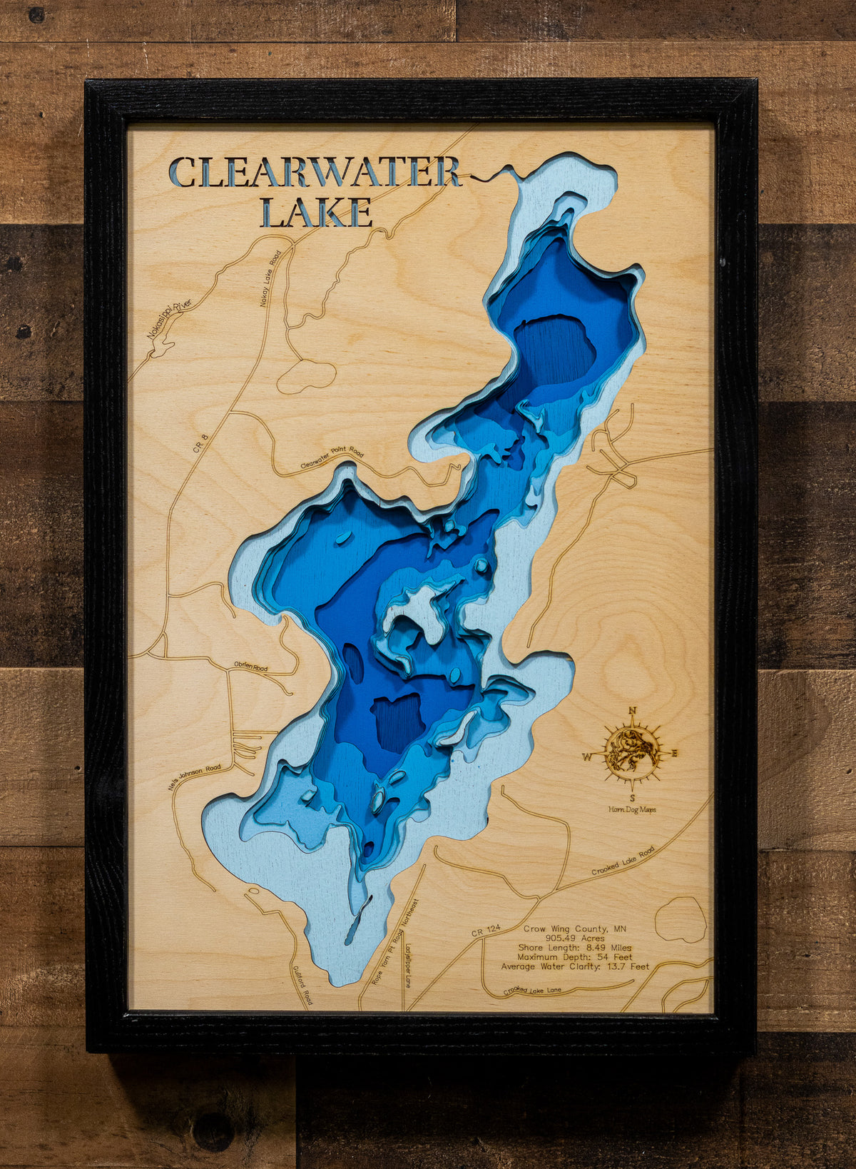 A framed wooden depth map of Clearwater Lake, featuring layered blue and light blue wood sections representing water depth, with contour lines and a black frame, mounted on a rustic dark wood plank wall.