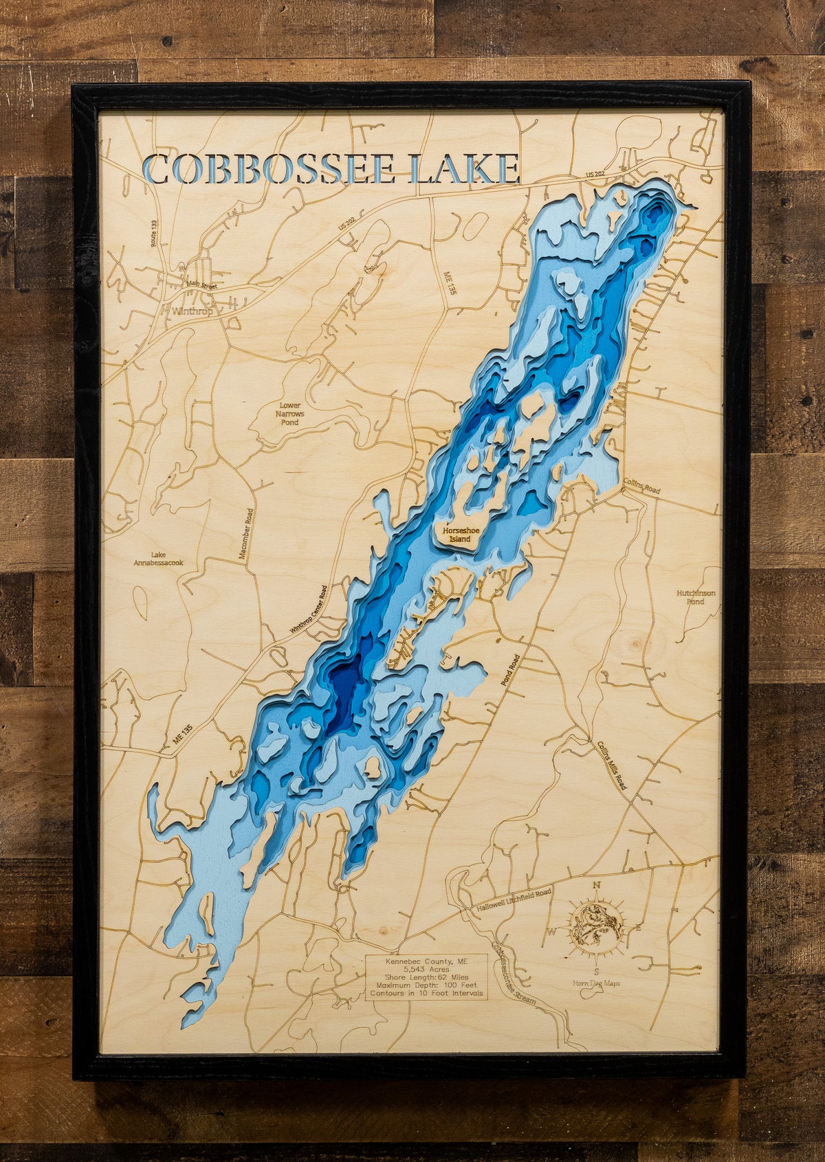 A framed wooden depth map of Cobbossee Lake, featuring blue laser-engraved water levels on a light natural wood background, enclosed in a black frame and mounted on a rustic dark wood plank wall.
