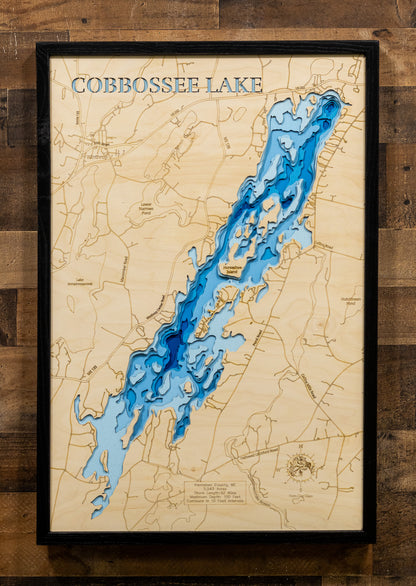 A framed wooden depth map of Cobbossee Lake, featuring blue laser-engraved water levels on a light natural wood background, enclosed in a black frame and mounted on a rustic dark wood plank wall.