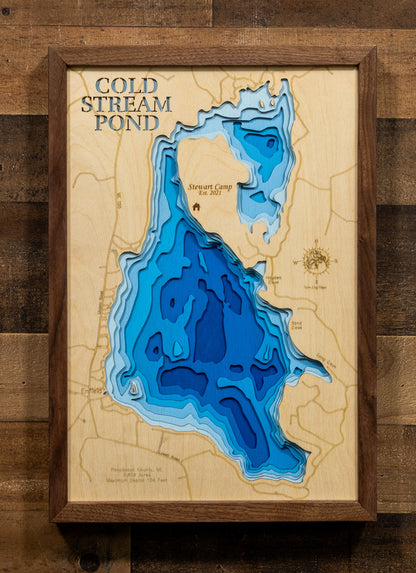A framed 3D layered wooden map of Cold Stream Pond, featuring deep blue and light blue concentric layers representing water depth, set against a light wood background with a dark brown wooden frame, mounted on a rustic wood plank wall.