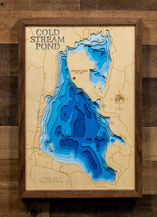 A framed 3D layered wooden map of Cold Stream Pond, featuring deep blue and light blue concentric layers representing water depth, set against a light wood background with a dark brown wooden frame, mounted on a rustic wood plank wall.