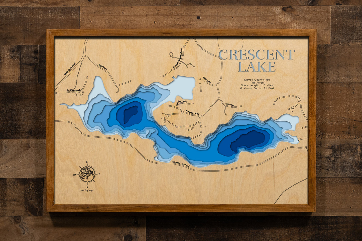 A beautifully detailed, laser-cut, multi-layered wooden topographical map of Crescent Lake in Carroll County, New Hampshire, showcasing the 148-acre lake's distinctive crescent moon shape with depth contours from the 10-foot average to the 21-foot maximum depth. The natural wood top layer frames 1.5 miles of shoreline including the Smith River inlet from Lake Wentworth and the dam outlet connecting to Lake Winnipesaukee, while graduated blue layers create an elegant visual depth gradient.
