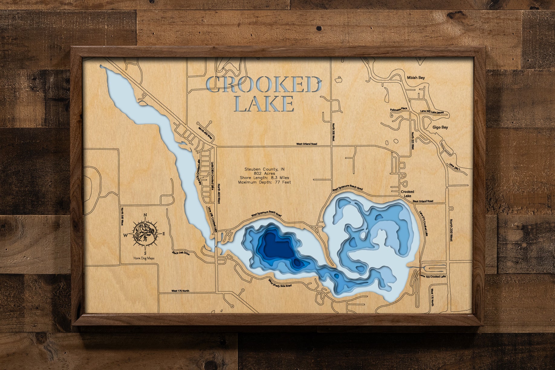 A laser-cut wooden map of Crooked Lake, Indiana, showcasing its unique three-basin structure across 802 acres. The intricate design features layered wood to illustrate the lake's 77-foot maximum depth and 8.3-mile shoreline. Key landmarks and surrounding Steuben County roads are precisely engraved. The natural wood grain highlights the North, Center, and West basins in a beautiful 3D topographic style.
